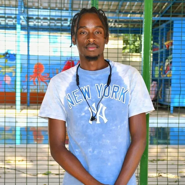 Daniel Kabiaru in a pale blue t-shirt, hands clasped, standing in front of a mesh fence with a painted sea mural behind him