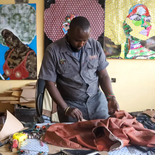 George Kamiti at the bench cutting leather, a row of his stitched portraits hanging on the wall behind him
