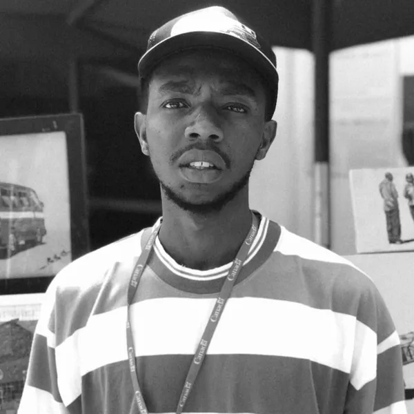 John Ruitha Maina in a cap and a striped shirt, standing in front of his stand with framed matatu paintings behind him