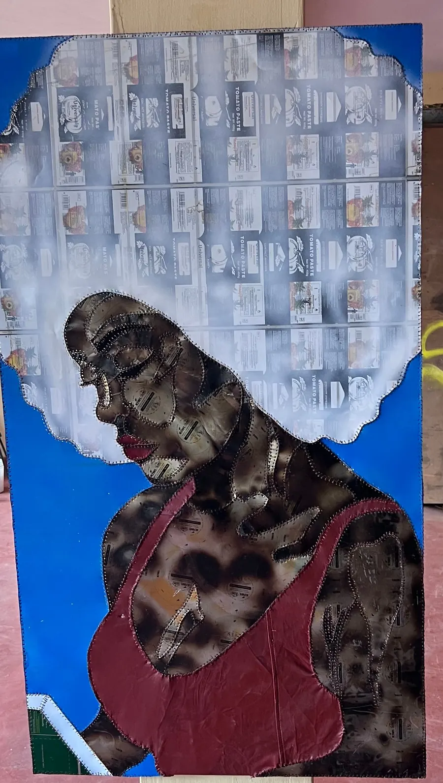 A stitched figure on a blue ground, the face and shoulders in scorched panels, a deep red leather bodice, and hair built from a grid of bleached tin labels