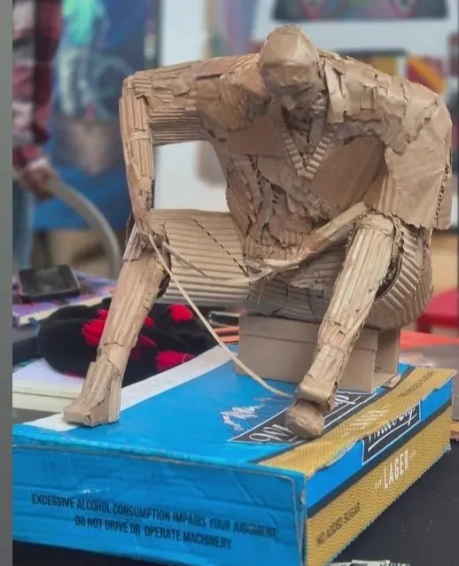 A seated figure built entirely from rolled and folded cardboard, elbows on knees, on a beer carton used as a plinth