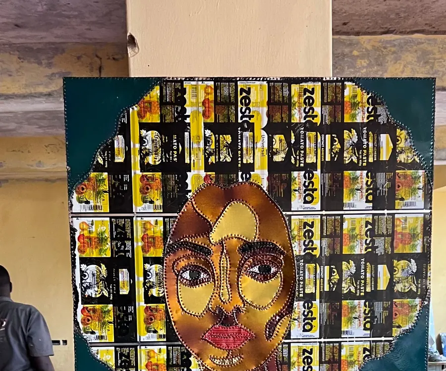 A stitched portrait on a dark teal ground, the face cut from warm sheet metal and the hair built from a grid of flattened Zesta tomato paste labels