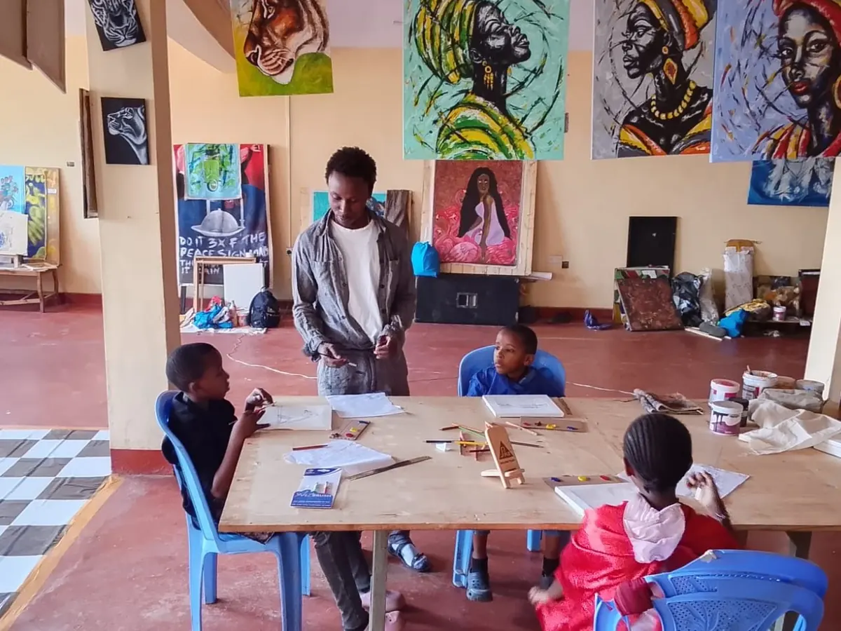 Dennis Bull Ndegwa standing at the long table while three children draw, under paintings hung from the studio ceiling